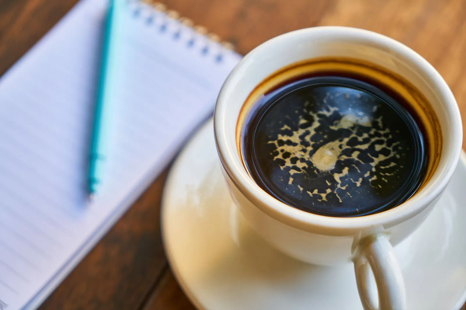 An open notebook beside a cup of coffee on a desk - the corner of an office where a shelf could sell books.
