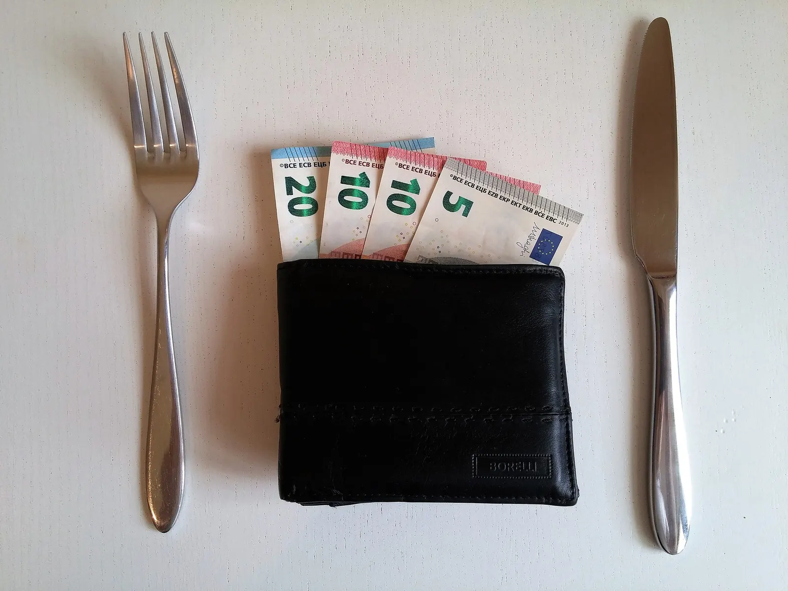 A wallet with banknotes set between a knife and fork on a white table - a book that earns its keep.