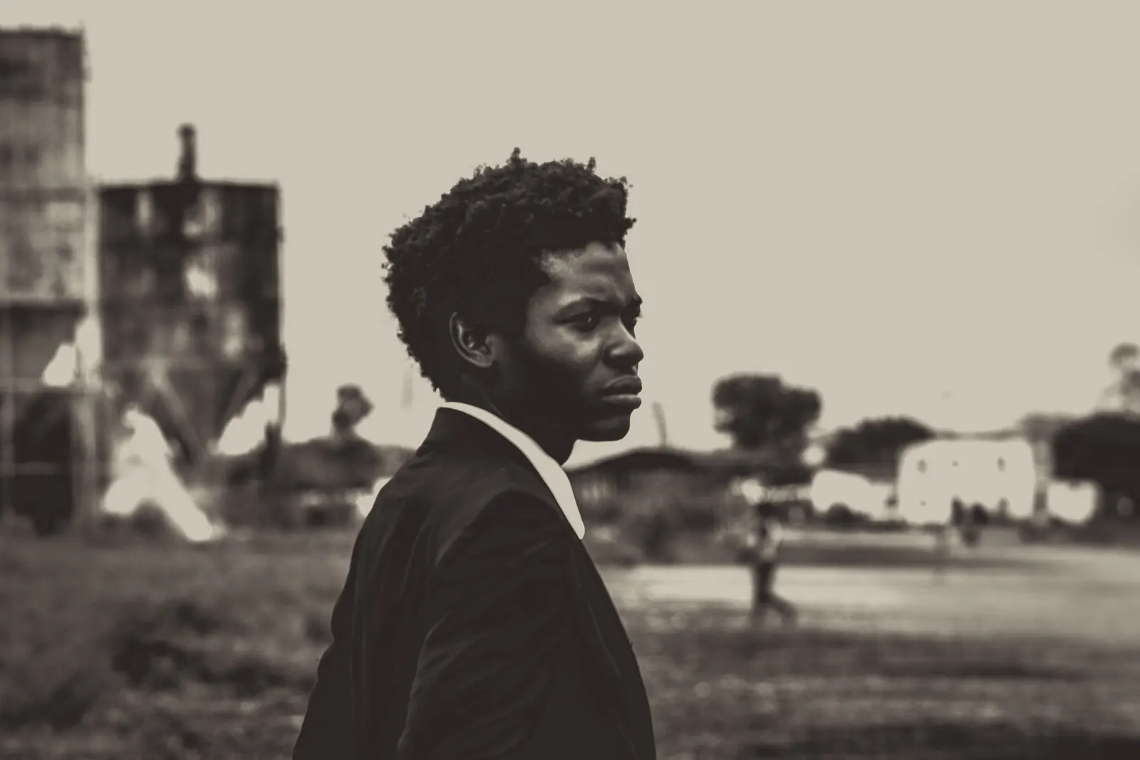 A writer in a dark suit stands alone in a sepia street of old buildings - the author before the book.